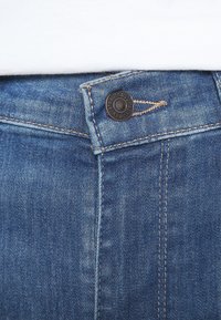 Jeans de mezclilla azules con cierre de botón y costuras contrastantes. Primer plano del área de la cintura con un remache de metal.