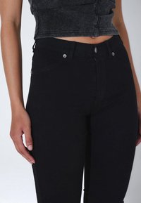 Femme portant un jean noir taille haute et un top court sans manches à côtes, avec un bras détendu le long du corps.