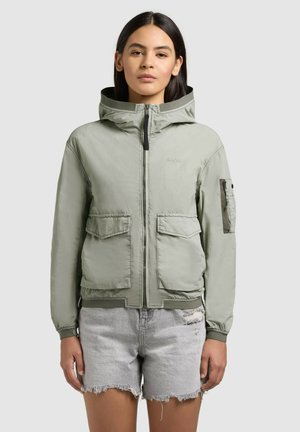 Vrouw die een lichtgroene capuchonjas met voorzakken draagt en lichtgrijze versleten denim shorts, staand tegen een effen achtergrond.