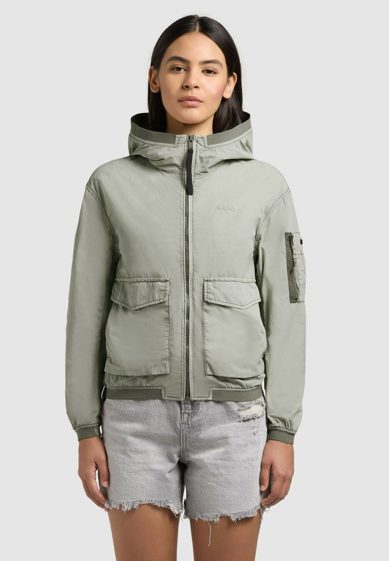Femme portant une veste à capuche vert clair avec des poches avant et un short en denim gris clair déchiré, debout devant un fond uni.