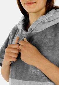 Grauer Frottee-Poncho mit Kapuze und Zugband, ausgestattet mit einem zweifarbigen Design und weicher Textur, von Händen in der Nähe des Halsauschnitts gehalten.