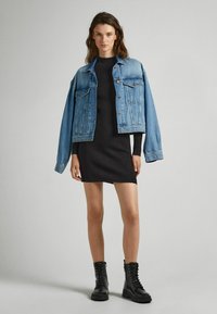 Giacca in denim con vestibilità rilassata, di colore azzurro, due tasche sul petto, abbinata a un vestito nero a coste e stivali da combattimento neri con lacci.