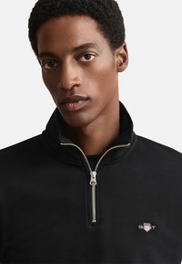 Sweatshirt preta com fecho de correr, feita de tecido macio, com gola alta, zipper prateado destacado e pequeno logotipo bordado no peito.