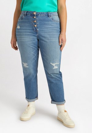 Blaue hochgeschnittene Jeans mit abgeriebenen Stellen und Fünf-Knopf-Verschluss, kombiniert mit weißen Sneakers und einem grünen Oberteil. Glatte Denim-Textur.