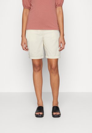 Person iført beige shorts med opkrøllede kanter, en støvet rosa kortærmet top og sorte åben-tå slip-on sandaler, stående på en ensfarvet baggrund.