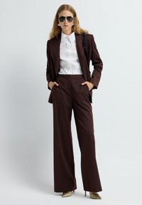 Mujer con traje de rayas marrón oscuro y pantalones de pierna ancha, camisa blanca con cuello, gafas de sol, tacones beige y bolso negro sobre fondo blanco.