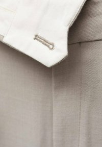 Guler de cămașă din bumbac bej deschis, cu cusături detaliate, având un accent unic la butonieră. Textură moale și design casual.