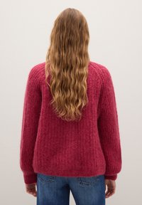 Lunghi capelli mossi di un marrone chiaro che scendono su un maglione rosso di maglia indossato con jeans blu, visti da dietro su uno sfondo bianco semplice.