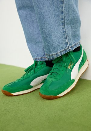Baskets vertes Puma Easy Ride avec bande blanche portées avec un jean bleu sur fond de tapis vert