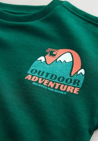 Le sweatshirt vert présente un design graphique avec des montagnes et un téléphérique. Le texte indique "AVENTURE EN EXTERIEUR" en lettres grasses et colorées.