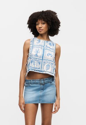 Młoda kobieta o kręconych włosach w bezrękawniku typu crop top z niebieskimi wzorami stempla i jasnoniebieskiej dżinsowej mini spódnicy z paskiem.