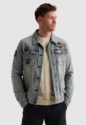 Mann trägt hellblaue Jeansjacke mit Aufnähern auf Brust und Ärmel, beigen Pullover und schwarze Hose, schaut nach rechts.