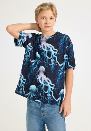 Lyshåret teenager iført en navyblå t-shirt med et mønster af lyseblå og lilla blæksprutter, kombineret med lyseblå jeans, stående med hånden bag hovedet.