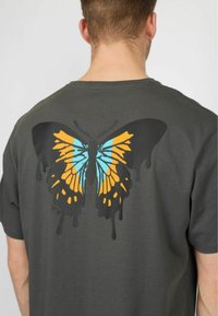 Grå t-shirt med et stort sommerfugleprint i sort, orange og blå med dryppende malerdetaljer på ryggen. Korte ærmer, rund hals.