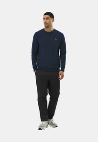 Felpa blu navy con logo rosso, collo a giro, polsini e orlo a coste, abbinata a pantaloni neri e scarpe da ginnastica grigio chiaro, in piedi su sfondo bianco.