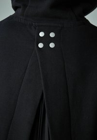 Čierny sveter s kapucňou, dvoma plochými skladmi na chrbte a štyrmi okrúhlymi striebornými cvokmi ako akcenty. Mäkšia textúra, neformálny dizajn.