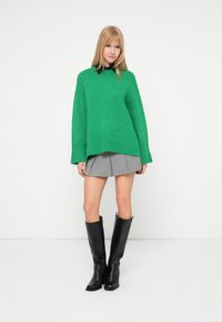 Πράσινη υπερμεγέθη πλεχτή ζακέτα με ραμμένο στρίφωμα, γκρι πλισέ μίνι φούστα και μαύρες ψηλές μποτάκια με τακούνι; λεία υφή και χαλαρός σχεδιασμός.