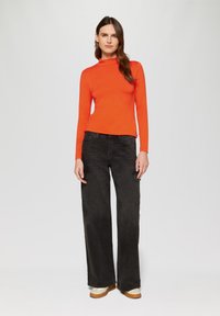 Rotes Langarmshirt mit Stehkragen, kombiniert mit schwarzen weit geschnittenen Cargo-Hosen mit Fronttaschen und hellen Schuhen.