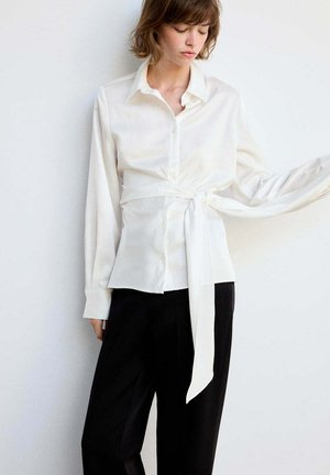 Femme portant une chemise blanche à manches longues avec une ceinture nouée à la taille et un pantalon noir, debout contre un mur blanc, regardant vers le bas.