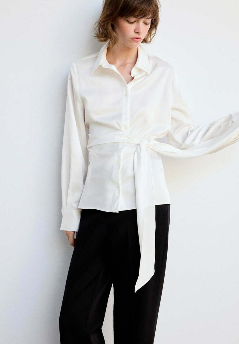 Femme portant une chemise blanche à manches longues avec une ceinture nouée à la taille et un pantalon noir, debout contre un mur blanc, regardant vers le bas.