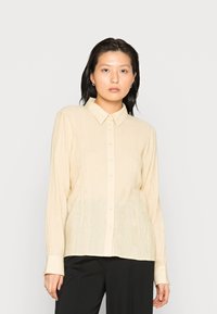 Camisa creme com botão, em tecido texturizado, mangas longas e colarinho clássico. Combinada com calças pretas.