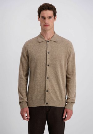 Mann trägt eine beige, knöpfbare Strickjacke mit Kragen und dunkle Hose, steht vor einem einfarbigen hellgrauen Hintergrund.