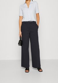 Donna che indossa una camicia azzurra chiara a maniche corte con bottoni, pantaloni navy a vita alta e a gamba larga, tacchi aperti neri e tiene una piccola borsa nera.