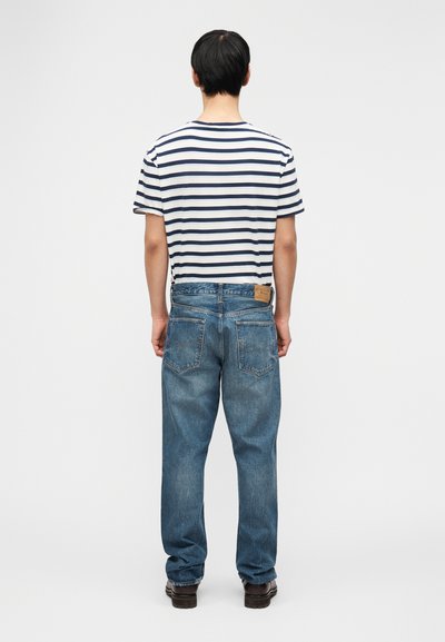 Čovjek stoji okrenut leđima, nosi bijelu majicu s plavim prugama, široke plave traperice i tamno smeđe cipele, na jednostavnoj pozadini.