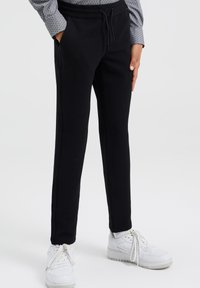 Pantaloni jogger neri con texture, tasche laterali, cintura elastica e cordoncino. Abbinati a scarpe da ginnastica bianche, che mettono in evidenza uno stile casual.