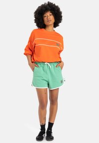 Hanorac portocaliu cu dungi albe și logo, asortat cu pantaloni scurți verzi cu borduri albe și buzunare. Încălțăminte neagră completează look-ul.
