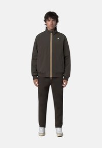 Giacca marrone con zip e colletto alto, strisce gialle e bianche a contrasto, polsini elasticizzati, abbinata a pantaloni marroni coordinati e sneakers bianche.