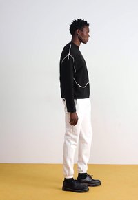 Sort sweater med hvide syninger, parret med hvide tapered bukser og sorte snørebåndssko på en gul baggrund.