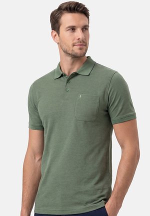 Uomo con polo verde a maniche corte con taschino sul petto, in piedi davanti a uno sfondo chiaro uniforme, che guarda di lato.