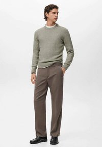 Hombre de pie, vistiendo un suéter de cuello redondo gris claro, pantalones marrones y zapatos de vestir negros, con las manos en los bolsillos, mirando hacia un lado sobre un fondo blanco.