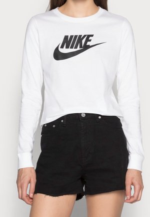 Femme portant un t-shirt blanc à manches longues Nike avec un logo noir et un short en jean noir taille haute avec bouton et poches visibles.