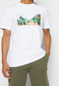 T-shirt bianco in cotone con una grafica colorata che ritrae paesaggi, con il testo "ellesse" sopra e "EST MCMLX" sotto.