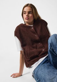 Joven sentada frente a un fondo blanco, vestida con chaleco sherpa marrón sobre camisa blanca y vaqueros azules, mirando hacia un lado.