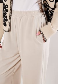 Vrouw in beige wijde broek met elastische tailleband en een crèmekleurige jas met zwarte geborduurde randen, hand in haar zak.