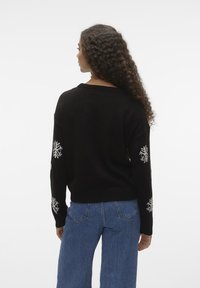 Svart strikkegenser med løs passform, med hvite snøfnuggmønstre på ermene. Kombinert med høyhalsede blå jeans.