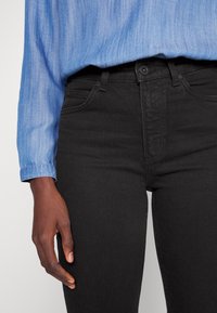 Jeans de mezclilla negra con corte ajustado, que cuentan con bolsillos frontales, pasadores para cinturón y cierre de botón. Combinados con una camisa de color azul claro.