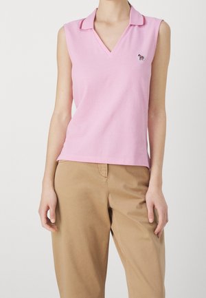 Różowa koszula bez rękawów z kołnierzykiem i małą naszywką zebry, noszona z beżowymi spodniami przez stojącą osobę na białym tle.