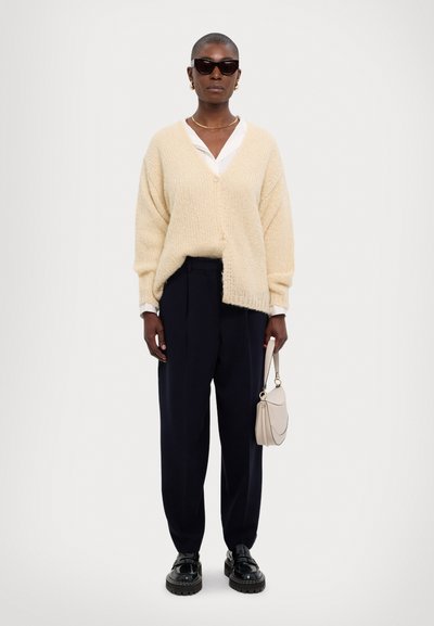 Cremefarbener Cardigan mit Knöpfen, weiße Bluse, dunkle Hose und schwarze Schuhe. Braune Handtasche mit geschwungener Form; Person trägt Sonnenbrille.