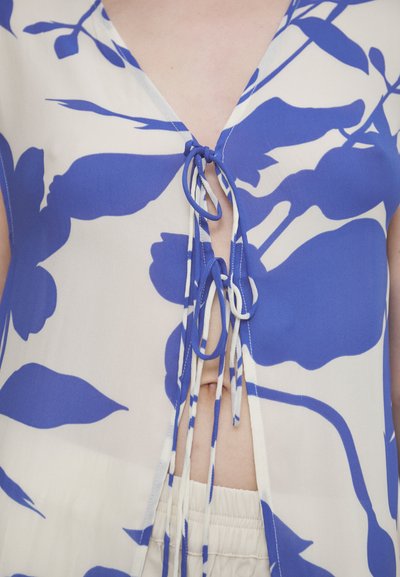 Bloemtop van doorschijnende stof met een blauw-wit patroon, voorzien van een diepe V-hals en striksluiting aan de voorkant. Lichte textuur, relaxte pasvorm.