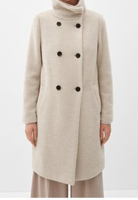 Cappotto in lana a spina di pesce beige con colletto alto, bottoni neri doppi, tasche laterali, indossato su pantaloni larghi marrone chiaro.