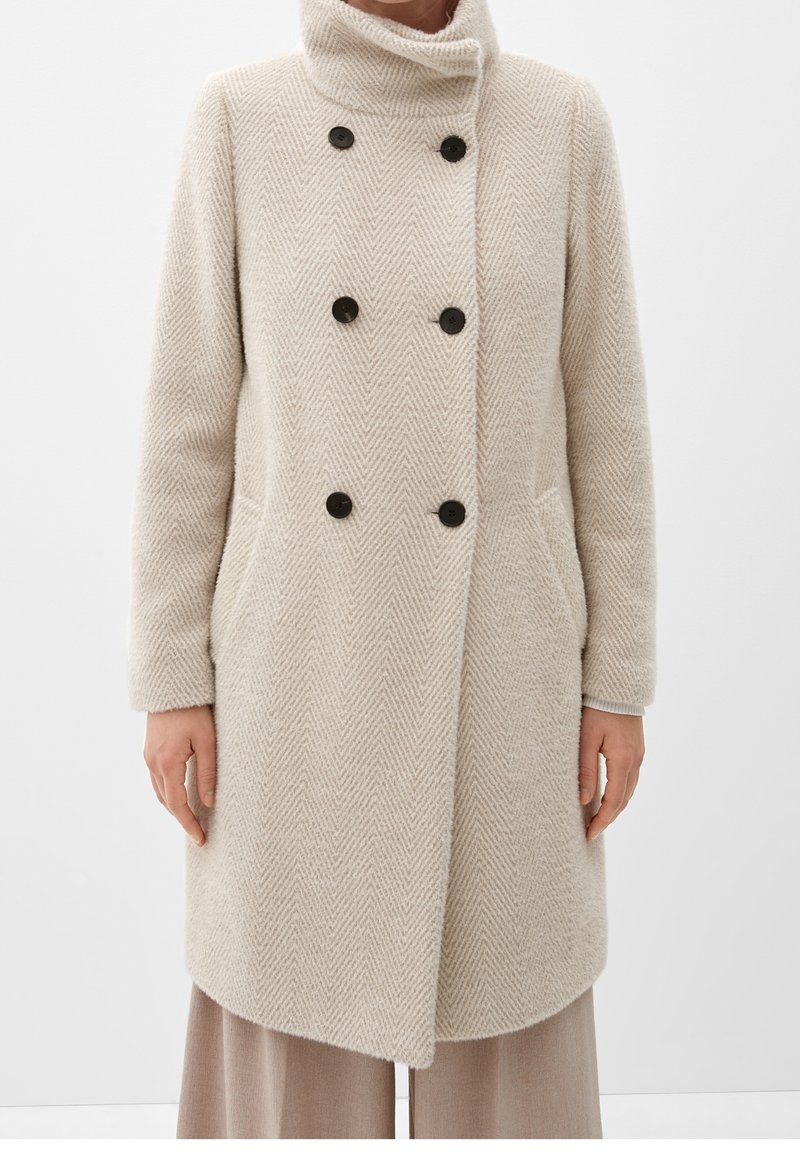 Cappotto in lana a spina di pesce beige con colletto alto, bottoni neri doppi, tasche laterali, indossato su pantaloni larghi marrone chiaro.