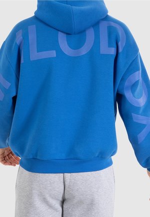 Personne portant un sweat à capuche bleu avec de grandes lettres violettes sur le dos et les manches, associée à un pantalon de survêtement gris clair.