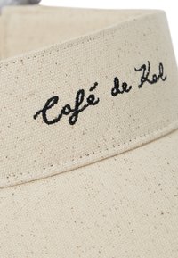 Kremfarget bomullslue med teksturert stoff, med svart brodert tekst "Café de Kol" på frontbåndet.