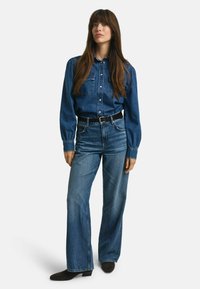 Blauwe denim shirt met een knoopsluiting aan de voorkant, gecombineerd met high-waisted blauwe jeans. Zwarte riem accentueert de taille, aangevuld met donker suède enkellaarsjes.