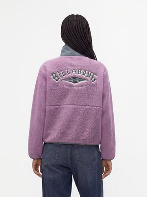 Personne portant une veste polaire violette avec le logo "Billabong" et un motif vague dans le dos, associée à un jean en denim bleu, vue de dos.