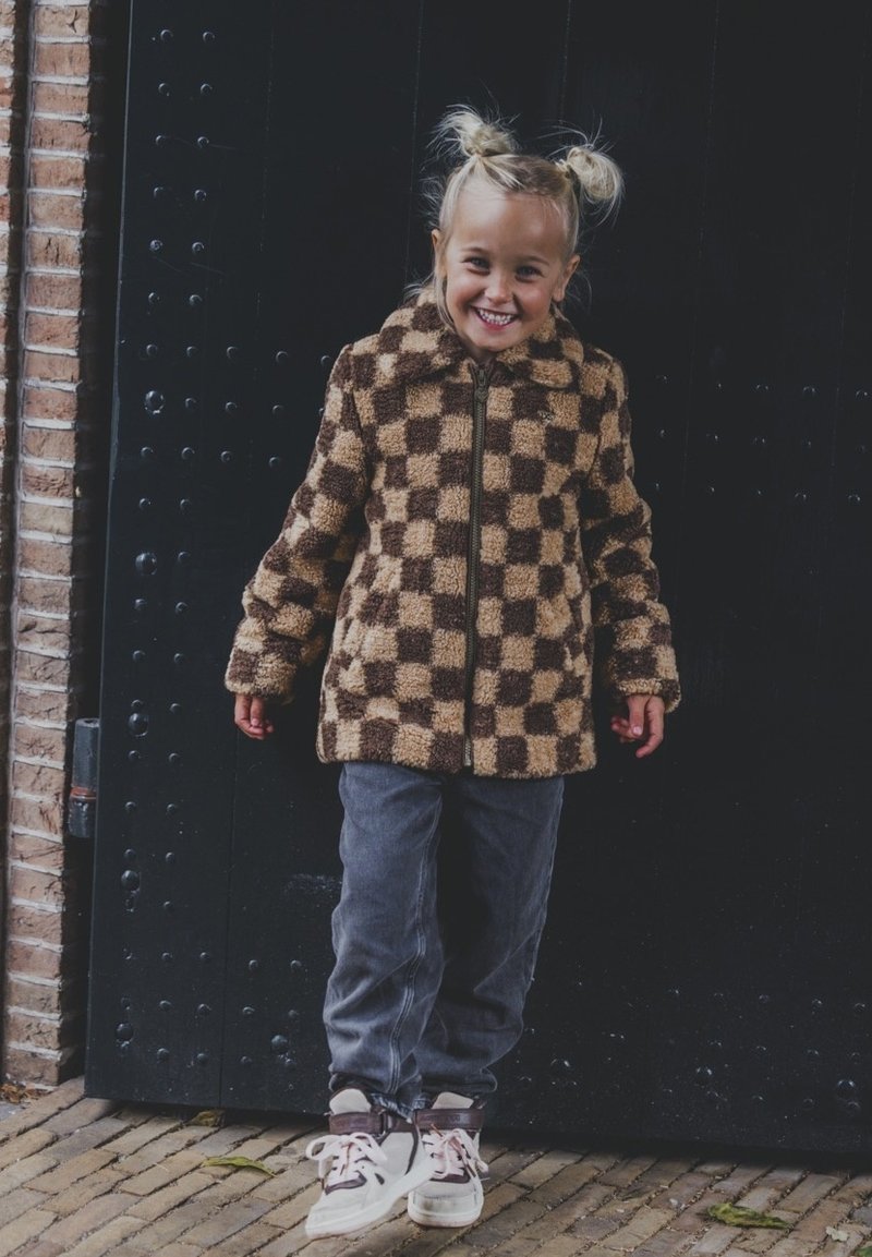 Bruine en beige geblokte fleece jack met een rits aan de voorkant, gedragen over grijze broek en witte sneakers. Gelaagd oppervlak, gezellige ontwerp.